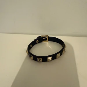 Valentino svart armband med nitar - Svart armband från Valentino i läder med guldiga pyramidformade nitar runt om. Justerbart spänne i guldton. Perfekt accessoar för att ge din look en edgy touch.