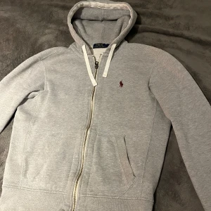 Grå hoodie från Polo Ralph Lauren - Väldigt eftertraktad och snygg ljusgrå hoodie från Polo Ralph Lauren med dragkedja, huva och vita snören. Klassisk broderad logga på bröstet och två fickor framtill. Perfekt för en chill och stilren look.