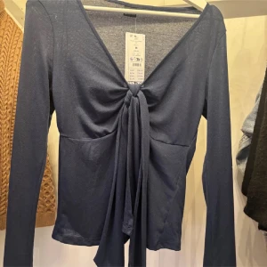 Mörkblå långärmad knytblus - Snygg mörkblå långärmad topp med djup v-ringning och knyt framtill. Toppens material är mjukt och lätt, perfekt för att skapa en chill men ändå stilren look. Passar dig som gillar trendiga detaljer och vill ha något som sticker ut.