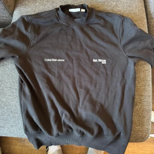 Svart sweatshirt från Calvin Klein Jeans - Svart sweatshirt från Calvin Klein Jeans med vit text på bröstet. Tröjan har ribbade muddar vid hals, ärmslut och nederkant. Enkel och stilren design som passar perfekt till jeans eller joggers.