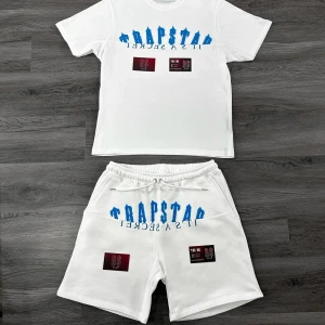 Vita Trapstar set med blå text - Snygga vit set från Trapstar med blå broderad logga och text framtill. Shortsen har elastisk midja med snörning och två patchar i rött och svart på benen. Perfekta för en chill och trendig look.