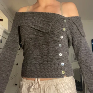 Grå offshoulder stickad tröja med knappar - Snygg grå stickad tröja med offshoulder-modell och snygga knappar längs ena sidan framtill. Tröjan har lång ärm och en lite croppad passform som ger en trendig vibe. Perfekt för dig som gillar en stilren look.