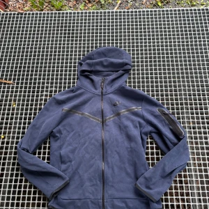 Blå Nike hoodiejacka med dragkedja - Snygg blå hoodiejacka från Nike med hel dragkedja framtill och huva. Jackan har en diskret logga på bröstet, reflekterande detaljer över bröstet och en praktisk ficka med dragkedja på ena ärmen. Perfekt för chill dagar eller när du är på språng.