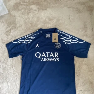 PSG fourth kit 24/25 - Blå PSG x Jordan fotbollströja med vita detaljer och vingar längs axlarna. Tryck med Qatar Airways på bröstet och Air Jordan-logga. Tillverkad i Dri-FIT-material som andas. Perfekt för dig som vill sticka ut på planen eller gatan.