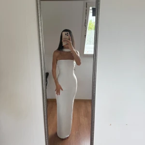 Vit axelbandslös långklänning - Supersnygg vit långklänning med axelbandslös design och hög slits bak. Klänningen har en tight passform och en stilren choker-detalj runt halsen. Perfekt för dig som vill sticka ut med en minimalistisk och elegant look. Använd en gång 