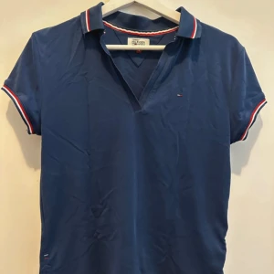 Blå pikétröja Tommy Hilfiger Denim - Snygg marinblå pikétröja från Tommy Hilfiger Denim med klassisk krage och korta ärmar. Tröjan har röd, vit och marinblå rand på krage och ärmslut samt liten logga på bröstet. Tillverkad i mjuk bomull, perfekt för en clean och sportig look.