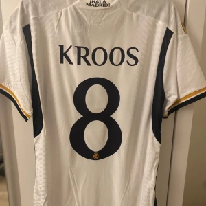 Real Madrid Kroos 8 Adidas fotbollströja - Snygg Real Madrid matchtröja med Kroos och nummer 8 på ryggen. Vit med guld- och marinblå detaljer, Adidas-logga och klubbmärke på bröstet. Slim fit-modell i lätt och ventilerande material, perfekt för fotbollsälskare. Helt ny bara att jag köpt in i fel storlek.