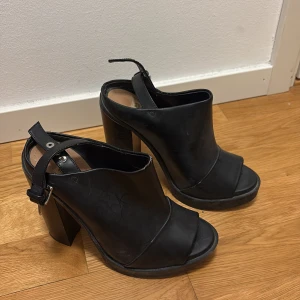 Svarta klackskor från Zara Trafaluc - Chunky svarta klackskor i skinnimitation från Zara Trafaluc. Skorna har öppen tå, bred klack och justerbar rem med silverdetaljer runt hälen. Perfekt för dig som gillar statement-heels med edgy vibe.