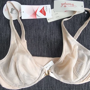 Beige mesh bh från Triumph 75B - Säljer en beige bh från Triumph med tunna, transparenta mesh-kupor och justerbara axelband. Bh:n har bygel och diskret guldfärgad detalj framtill. Perfekt för dig som gillar en stilren och enkel look.