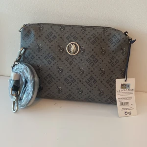 Grå axelväska från U.S. Polo Assn. - Snygg grå axelväska från U.S. Polo Assn. med mörkgrått mönster och logga framtill. Väskan har justerbar axelrem, dragkedja upptill och detaljer i silverfärgad metall. Tillverkad i slitstark syntet med klassiskt tryck och praktiskt innerfack. Ny pris 900kr