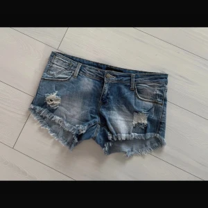 Blå slitna jeansshorts med fransar - Säljer ett par blå jeansshorts med slitna detaljer och fransig kant. Shortsen har klassisk femficksmodell, låg midja och coola trasiga partier framtill för en avslappnad vibe. Perfekta för sommardagar när du vill ha en chill och trendig look.