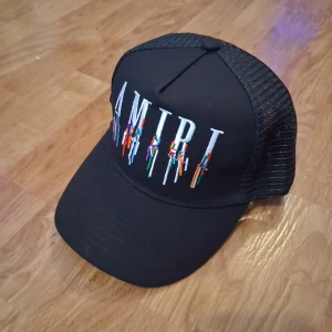 Svart keps från Amiri med brodyr - Svart truckerkeps från Amiri med justerbar snapback och mesh baktill. Framsidan har stor broderad AMIRI-logga i vitt med färgglada detaljer. Kepsen är tillverkad i bomull och elastan för skön passform och cool look.