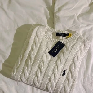 Polo Ralph Lauren sweatshirt  - Vit kabelstickad tröja från Polo Ralph Lauren med klassisk rund halsringning och broderad marinblå logga på bröstet. Tröjan har lång ärm och är tillverkad i mjuk bomull, perfekt för en clean och stilren look.