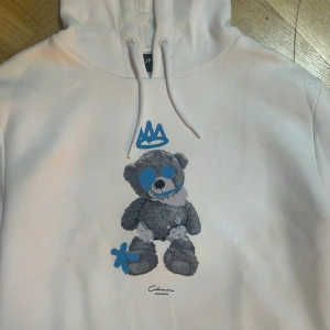 Vit hoodie med nalletryck från Closure - Vit hoodie från Closure London med ett stort tryck av en grå nalle med blå detaljer och krona på bröstet. Tröjan har huva med snörning och en stor magficka. Perfekt för dig som gillar streetstyle och unika prints.
