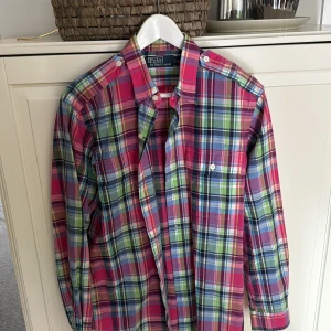 Rutig skjorta från Polo Ralph Lauren - Färgglad rutig skjorta från Polo Ralph Lauren i bomull. Skjortan har lång ärm, klassisk krage och knäppning framtill. Färgerna går i rosa, blått, grönt och vitt, vilket ger en riktigt fräsch vibe. Perfekt för dig som gillar att sticka ut lite extra.