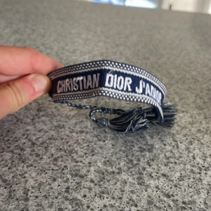 Christian Dior J'Adior armband blå - Snyggt vävt armband från Christian Dior med texten 'J'Adior' i vitt på marinblå botten. Armbandet har justerbara snören med tofsar i matchande färger. Perfekt accessoar för att lyfta din outfit med en lyxig touch.