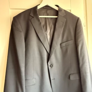 Svart kavaj från Dressmann, stl 58 - Klassisk svart kavaj från Dressmann i storlek 58 Short. Enkelknäppt modell med två fickor fram och en bröstficka. Snyggt snitt och tidlös design, perfekt till skjorta och byxor. Materialet känns slätt och stilrent.