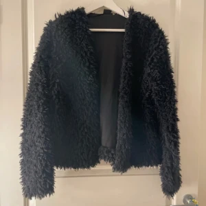 Svart fluffig teddyjacka - Säljer en svart teddyjacka med långärmad design och oversized känsla. Jackan är supermjuk och har en fluffig pälslook som ger en cool vibe. Perfekt för dig som vill ha en mysig och trendig jacka till höst eller vintern.