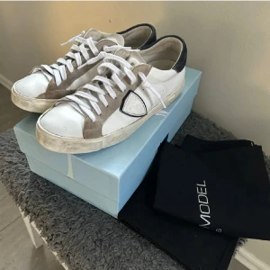 Philippe Model vita och beige sneakers - Snygga sneakers från Philippe Model i vitt skinn med beige mockadetaljer och mörkblå häl. Klassisk låg modell med vit sula och vita snören. Skorna har en ikonisk sköld på sidan och är perfekta för en avslappnad men trendig stil.