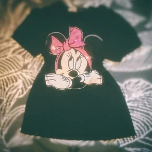 Svart Minnie Mouse t-shirt med paljetter - Svart t-shirt med korta ärmar och stor tryckt bild av Minnie Mouse på framsidan. Minnies rosett är rosa och dekorerad med paljetter som ger extra bling. Perfekt för dig som gillar Disney och vill sticka ut med en söt och lekfull stil.