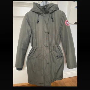 Grön parkas jacka från Canada Goose S - Snygg grön parkajacka från Canada Goose med huva, dragsko i midjan och klassisk logga på ärmen. Jackan har långa ärmar, stora fickor framtill och dragkedja. Perfekt för kalla dagar när du vill hålla stilen och värmen.