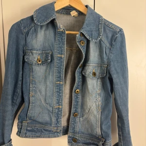 Blå jeansjacka från Vero Moda - Klassisk blå jeansjacka från Vero Moda med snygga bronsfärgade knappar och två bröstfickor med lock. Jackan har en markerad krage, dekorativa sömmar och justerbara spännen i midjan. Skriv vid frågor!💞