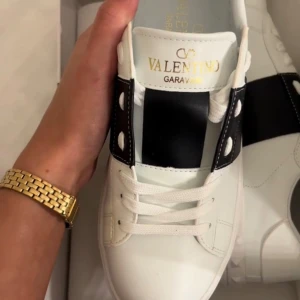 A Kopior Valentino Garavani Open sneakers vit/svart - Snygga Valentino Garavani Open sneakers i vitt skinn med bred svart läderdetalj över vristen och ikoniska nitar på sulan. Klassisk rund tå, vita snören och guldfärgad logga på plösen. Perfekta för dig som gillar lyxiga och trendiga sneakers.