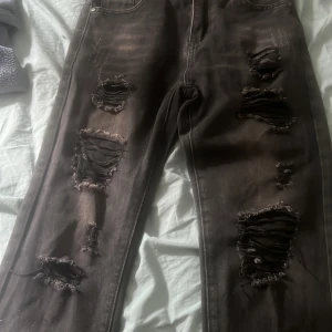 Svarta ripped jeans med dragkedja - Säljer ett par svarta jeans och trasiga detaljer på benen för en edgy look. Jeansen har dragkedjegylf och coolt mönstrat innerfoder i midjan. Perfekta för dig som gillar streetstyle och vill sticka ut.