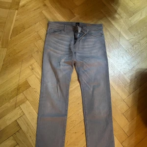 Gråa jeansbyxor från Hugo Boss Maine - Säljer ett par gråa Hugo Boss Maine jeans med regular fit. Klassisk femficksmodell med orangea sömmar och snyggt tvättad look. Byxorna har rak passform och är tillverkade i mjukt bomullstyg med en hint av stretch för extra komfort.