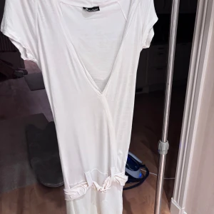 Vit omlottklänning med huva - Supersöt vit kortklänning/tunika från Urban Angel med omlott-skärning, djup v-ringning och korta ärmar. Plagget har en huva och ett brett bälte med tygspännen i midjan som ger en cool detalj. Mjuk och lätt i materialet, perfekt för sommarens varma dagar.