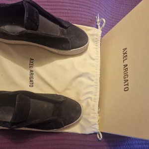 Svarta sneakers i mocka från Axel Arigato - Stilrena svarta sneakers från Axel Arigato i mjuk mocka med vit sula. Skorna har minimalistisk design utan snörning och en bred elastisk rem över vristen för enkel påtagning. Perfekta för dig som gillar en clean och modern look.