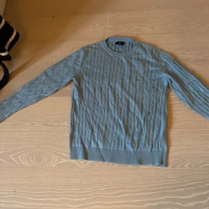 Ljusblå kabelstickad tröja från Gant - Snygg ljusblå kabelstickad tröja från Gant med rund hals och diskret broderad logga på bröstet. Tröjan har långa ärmar och ribbade muddar vid ärmslut och nederkant. Perfekt för dig som gillar klassisk och stilren look.