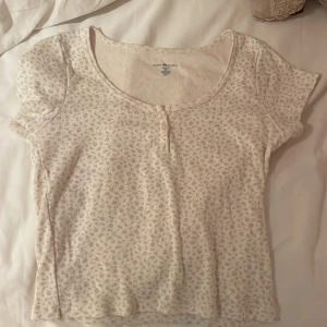 Blommig topp från Brandy Melville - Supersöt ljusbeige topp från Brandy Melville med små blommor i mönstret. Modellen har korta ärmar, rundad halsringning och två knappar framtill. Tillverkad i mjuk bomull 