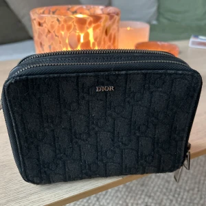 Svart Dior väska - Säljer en svart väska från Dior med diskret mönster och dubbla dragkedjor. Väskan har en stilren, rektangulär form och små metall-detaljer. Perfekt för dig som gillar klassisk design med en twist. (Tyvärr saknas axelremmen där av priset)
