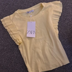 Gul ribbad topp med volangärm Zara - Supersöt ljusgul ribbad topp från Zara med volangdetaljer på ärmarna. Toppen har rund halsringning och är tillverkad i mjuk bomull, perfekt för en fräsch och somrig look. Säljs med prislapp kvar!