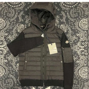 Moncler cardigan - Snygg svart pufferjacka från Moncler med stickade ärmar och quiltad kropp. Jackan har huva, dragkedja framtill och en cool ficka på ärmen med Moncler-logga. Perfekt för dig som vill ha en jacka med både street och lyxkänsla.