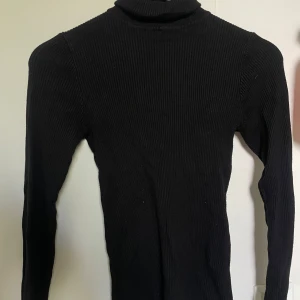 Svart ribbad polotröja från Amisu - Säljer en svart ribbad polotröja från Amisu i storlek S. Tröjan är långärmad och har en tight passform med hög krage. Perfekt att styla med jeans eller kjol för en clean look. Materialet är stickat och känns mjukt mot huden.