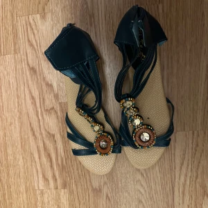 Sandaler med pärldetaljer och kilklack - Unika sandaler med mörkgröna remmar och snygga pärldetaljer i guld och trä. Sulan har flätad look och skorna har öppen tå samt kilklack. Perfekta för sommaren och ger en boho-vibe till din outfit.