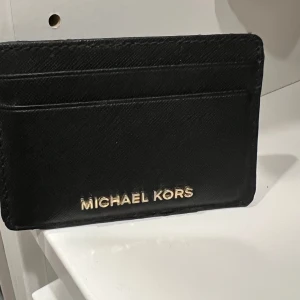 Svart korthållare från Michael Kors - Snygg svart korthållare från Michael Kors i stilrent skinn med guldiga bokstäver framtill. Perfekt för att hålla koll på dina kort och cash utan att ta plats. Enkel och clean design som passar i alla fickor eller väskor. Mindre slitage ovanpå kanten som visas på bilden. 