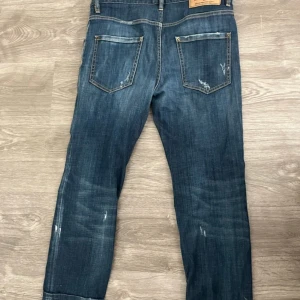 Dsquardjeans med slitningar med uppvikt nertill - Säljer ett par Dsquard jeans i mörkblå tvätt med slitna detaljer och fransade benslut. Jeansen har raka ben, fem fickor och tydliga slitningar för en cool, avslappnad look. Perfekta för dig som gillar vintage vibe och streetstyle. Köpta för 1200kr på Nylins & Skeppsteds.