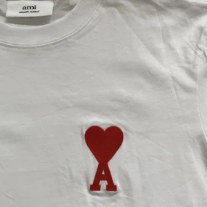 Vit t-shirt från Ami med rött hjärta - Vit t-shirt från Ami med klassisk passform och rund hals. På bröstet finns ett broderat rött hjärta med bokstaven A. Tillverkad i mjuk bomull, perfekt för en clean och stilren look.