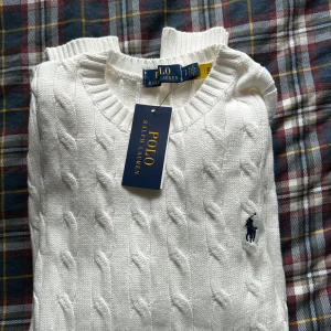 Polo Ralph Lauren tröja - Vit kabelstickad tröja från Polo Ralph Lauren med klassisk rund halsringning och broderad marinblå logga på bröstet. Tröjan har ribbade muddar och är långärmad, perfekt för en clean och stilren look.