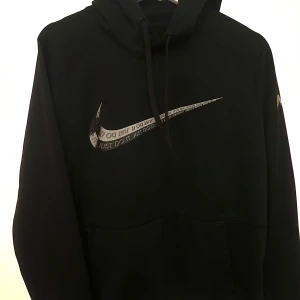 Svart Nike Therma-Fit hoodie M - Svart Nike Therma-Fit hoodie med stor silvrig Swoosh och texten 'Just Do It' på bröstet. Tröjan har huva med snörning och en stor magficka. Perfekt för dig som gillar sportig och clean stil.