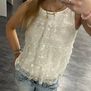Vit blommig spetstopp - Supersöt vit blus från H&M med broderade blommor och volangdetaljer. Ärmlös modell med rund halsringning och dubbla lager av tunn, transparent mesh.