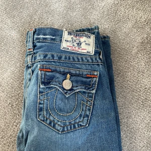 True Religion blå raka jeans - Säljer ett par klassiska blå jeans från True Religion med raka ben och ikonisk broderad bakficka. Jeansen har orangea detaljer vid sömmarna och silverfärgade knappar. Tillverkade i slitstarkt denim med snygg tvättad look.