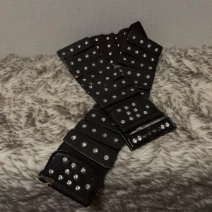 Svartbrunt höftbälte med nitar och strass - Brett midjebäste med strass och knäppning med dubbla hakar.  Fyra strasspärlor fattas vid spännet. Strasspärlorna sitter på ”plattor” med mjukt resårband under för skön passform.  Bältet är väl använt och har många år på nacken så skinnet flagnar väldigt lätt.   Längd ca 78 cm Bredd 7,5 cm