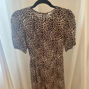 Leopardmönstrad klänning med öppen rygg från NA-KD storlek S - Snygg kortklänning från NA-KD med leopardmönster i storlek 34. Klänningen har puffiga korta ärmar och en öppen rygg. Figurnära upptill och lätt utsvängd med volangkant nertill.