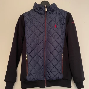 Quiltad blå jacka från Polo Ralph Lauren - Snygg quiltad jacka från Polo Ralph Lauren i mörkblått med svarta ärmar. Jackan har dragkedja framtill, två fickor med dragkedja och broderad röd logga på bröstet. Perfekt för dig som vill ha en sportig och stilren look.
