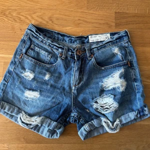Blå slitna jeansshorts Circle of Trust - Snygga blå jeansshorts från Circle of Trust med slitna detaljer och upprullade benslut. Klassisk femficksmodell med knäppning fram och coola rivna partier både fram och bak. Perfekta för en avslappnad sommarstil.