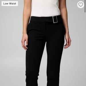 Svarta lågmidjade bootcut kostymbyxor - Stilrena svarta kostymbyxor med låg midja och bootcut ben. Byxorna har en bred linning med en snygg silverfärgad spänne och raka pressveck längs benen. Perfekta för en clean och trendig look. Går ända ner till fötterna på mig som är 168cm. 